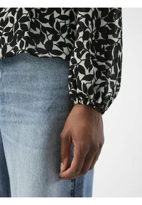 Blouse à motifs de feuilles en noir et blanc avec des manches bouffantes et des poignets élastiques, associée à un jean en denim bleu clair. Tissu doux et texturé visible.