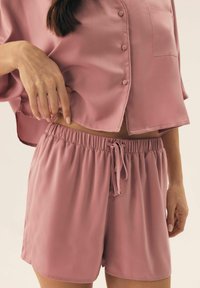 Różowy satynowy zestaw loungewear składający się z koszuli zapinanej na guziki oraz szortów z elastycznym paskiem i sznurkiem, o gładkiej teksturze i luźnym kroju.