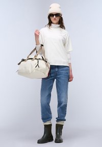 Beige sporttas met bruine banden, gecombineerd met een witte langemouwtop, blauwe jeans en zwarte rubberen laarzen met een gestructureerd oppervlak.