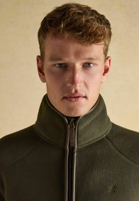 Zelená flísová bunda s vysokým golierom a zipsovým zapínaním. Obsahuje logo na ľavej strane a hladký, textúrovaný povrch.