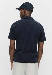 Marineblauwe short-sleeve shirt met een kraag, gestructureerde stof en naaddetails aan de achterkant, gedragen met lichtgekleurde broek en een bijpassende pet.