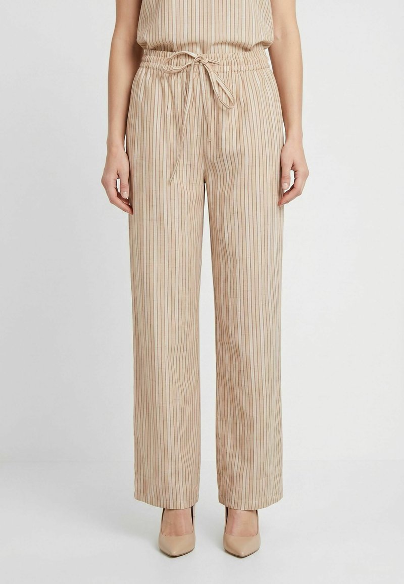 Pantalon large beige à rayures verticales avec une taille à cordon, porté avec des chaussures à talons beiges sur un fond uni.