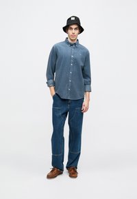 Košeľa s dlhým rukávom v tmavomodrej bavlne so jemným vertikálnym pruhovaním, skombinovaná s širokými modrými džínsami a hnedými člunkovými topánkami.