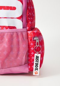 Ροζ σακίδιο με τσέπη από υφασμένο πλέγμα, μεγάλο λευκό λογότυπο, κυκλικά σχέδια και ετικέτα που γράφει "ΑΠΛΑ ΚΑΝΕ ΤΟ". Ορατή λεπτομέρεια από φερμουάρ.