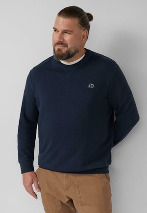 Marineblauwe sweatshirt van zachte stof met een ronde hals, voorzien van een klein logo-opject op de borst en geribde manchetten. Gestyleerd met beige broeken.