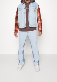 Gilet di denim azzurro chiaro con maniche a quadri, felpa marrone e jeans di denim chiaro con pannelli laterali scuri. Accompagnato da sneakers grigie.