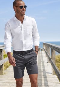 Camicia bianca di lino con maniche lunghe e taschino sul petto, abbinata a pantaloni corti grigio antracite. Cintura intrecciata nera con una fibbia elegante. Occhiali da sole indossati.