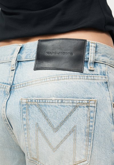 Ανοιχτό μπλε τζιν τζιν με κεντημένη λεπτομέρεια "M" στην τσέπη και μαύρο δερμάτινο λογότυπο Marc Jacobs στη μέση της μέσης, φορεμένο με μαύρο τοπ.