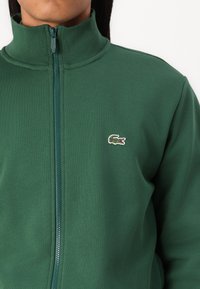 Sudadera verde con cremallera y cuello alto, confeccionada en tejido liso. Presenta un pequeño logo de cocodrilo bordado en el pecho izquierdo.
