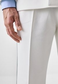 Beli teksturirani hlače s prilegajočim dizajnom in vidnim šivom, v kombinaciji s svetlo modro srajco. Roka se opira na material.