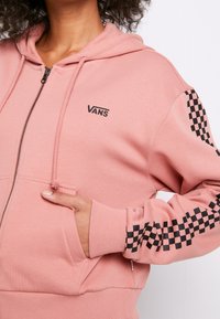 Persona che indossa una felpa con zip rosa Vans, con un motivo a scacchi neri sulle maniche, con una mano in tasca, mostrata dalla mascella alla vita.