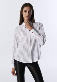 Camisa blanca de botones con finas rayas verticales negras; presenta un acento de corazón rojo en el puño. Combinada con leggings negros.
