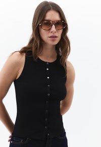 Top sans manches noir à côtes avec un col rond, fermeture à boutons sur le devant et coupe ajustée. Porté avec des lunettes de soleil à motifs et un jeans foncé.
