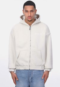 Hoodie zippé gris clair avec poche kangourou, poignets élastiques et capuche à cordon. Présente un logo subtil sur la poitrine. Tissu doux et texturé.