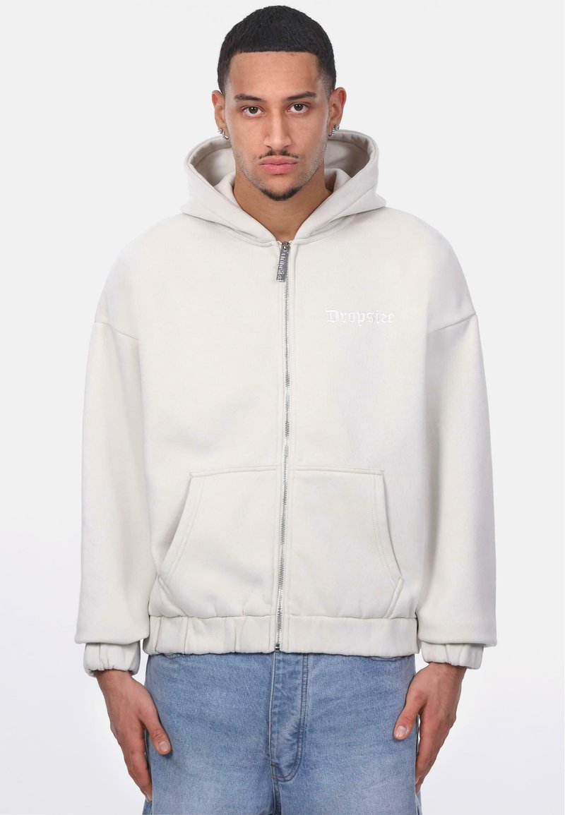 Hoodie zippé gris clair avec poche kangourou, poignets élastiques et capuche à cordon. Présente un logo subtil sur la poitrine. Tissu doux et texturé.