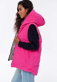 Růžový kordurový vesta s velkou kapucí, s podšívkou s leopardím potiskem, bočními kapsami a černým zipem. Stylizováno přes tmavý svetr.