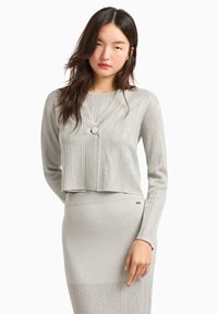 Cardigan corto grigio chiaro con scollo a V, texture a costine e un bottone decorativo. Indossato sopra una gonna abbinata, anch'essa grigio chiaro.