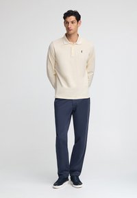 Kremowa koszula polo z długim rękawem i kołnierzykiem, z czarnym logo na piersi. Połączona z granatowymi spodniami i granatowymi trampkami.