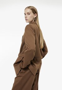 Brauner maßgeschneiderter Blazer mit lockerer Passform, minimalistischem Design, Ein-Knopf-Verschluss und zwei seitlichen Taschen. Glatter Stoff mit leichter Textur.