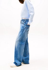 Person som bär vida blå jeans, vita sneakers och en ljusblå kortärmad sweatshirt med händerna i fickorna, stående på en vit bakgrund.