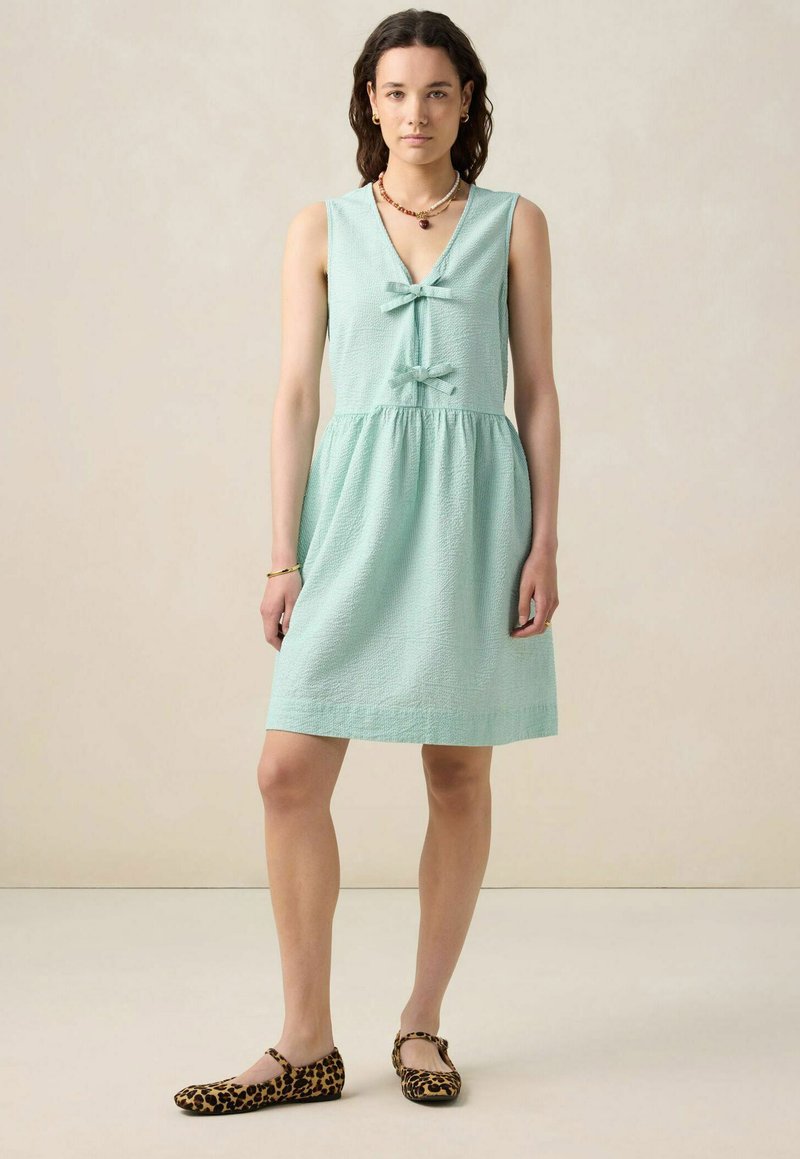 Robe sans manches vert menthe en tissu texturé, avec un décolleté en V, deux accents en nœud et une taille froncée, portée avec des ballerines à imprimé léopard.