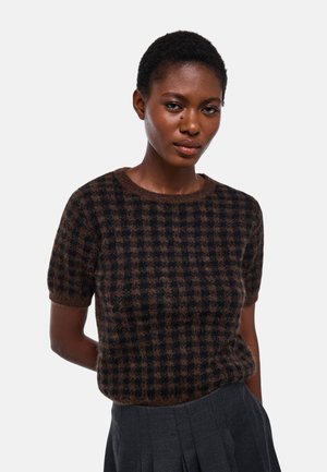 Femme aux cheveux courts portant un pull à manches courtes à carreaux marron et noir en tissu duveteux et une jupe plissée foncée sur un fond uni.