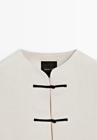 Hvid beklædning med rund halsudskæring med to sorte vandrette låsekroge og en synlig sort mærkeseddel indeni.