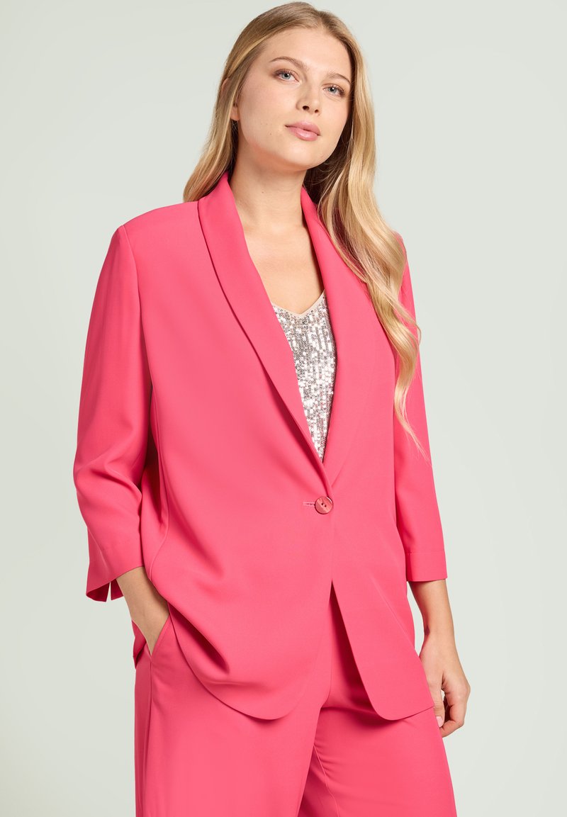 Frau mit langen blonden Haaren, die ein leuchtend pinkes Blazer und passende Hose über einem silbernen Pailletten-Top trägt, Hände in den Taschen, neutraler Hintergrund.