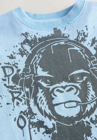 Gaiši zils jakas krekls ar lielu pelēka goruļa grafiku, kas valkā austiņas un ir ar traipu detaļām, apaļu kakla izgriezumu un ribotām aprocēm.