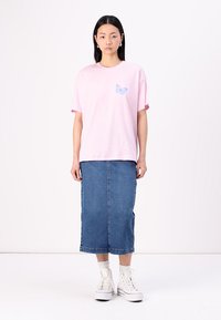 Pink cotton t-shirt with a blue butterfly print on the left chest, paired with a denim pencil skirt. Model wears white sneakers.