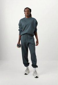 Ciemnoturkusowa bluza i dopasowane dresy, oba wykonane z miękkiego materiału. Spodnie mają elastyczny pas, modelka trzyma butelkę wody.