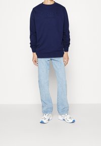 Persona che indossa un felpa navy blue di Lyle & Scott, jeans dritti azzurri e sneakers bianche con accenti blu, in piedi contro uno sfondo bianco.