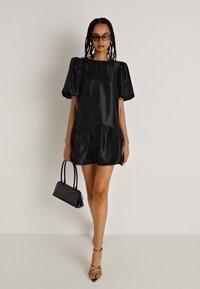 Μαύρο μίνι φόρεμα από σατέν με φουσκωτά μανίκια, ριγωτό τελείωμα και τετράγωνη λαιμόκοψη. Συνδυασμένο με μαύρη τσάντα και μπεζ ψηλοτάκουνα σανδάλια.