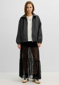Chaqueta bomber de mezcla de lana gris con cierre de cremallera, superpuesta sobre una camisa de algodón blanca y una falda maxi de encaje negro transparente. Zapatillas blancas con detalles en negro.