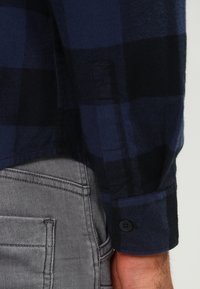 Chemise en flanelle à carreaux bleu marine et noir avec des poignets à boutons, associée à un pantalon en denim gris doté de poches latérales et de détails de couture visibles.