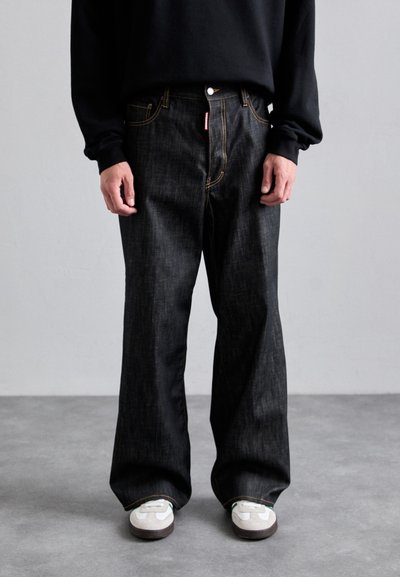 Person wearing black wide-leg jeans with visible orange stitching and white sneakers, standing on gray floor against plain light wall.