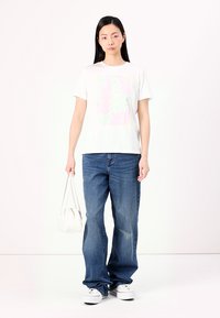 Witte korte mouwen t-shirt met een pastel bloemenprint, gecombineerd met losse blauwe jeans en witte sneakers. Model houdt een kleine witte handtas vast.
