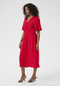 Red midi dress with short, wide sleeves and a knotted waist detail. Made from textured fabric, paired with black strappy heels.
