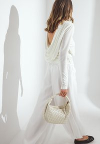 Weißes Oberteil mit Drapierung und Rückenausschnitt, kombiniert mit lockeren, fließenden weißen Hosen. Geflochtene hellbeige Handtasche mit rundem Henkel, schwarze flache Schuhe.