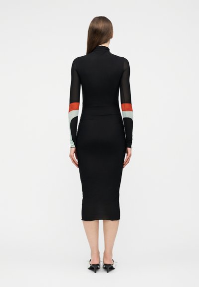 Nő, aki egy testhezálló fekete midi ruhát visel hosszú ujjakkal, amely piros, fehér és fekete színblokkokat tartalmaz, háttal állva a kamerának.