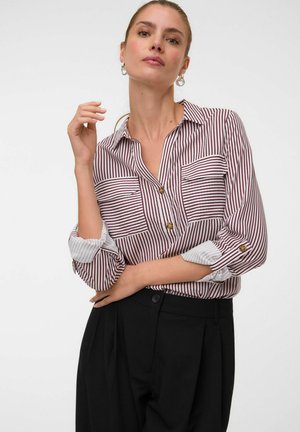 Femme portant une chemise à rayures verticales rouges et blanches à manches longues avec poignets retroussés et un pantalon noir taille haute.