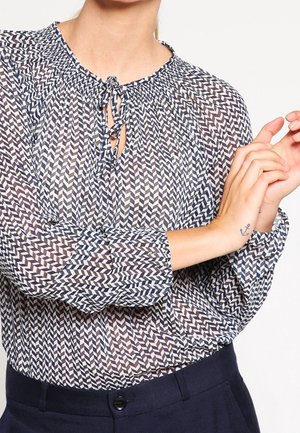Femme portant un chemisier à manches longues, à motifs, avec un col à nouer, un pantalon foncé, montrant un petit tatouage d'ancre sur son poignet.