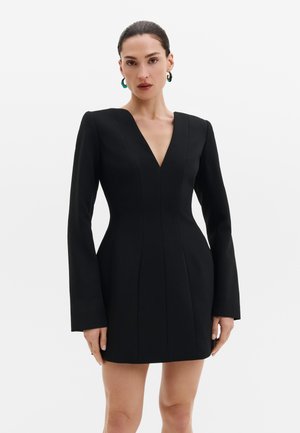Femme aux cheveux foncés portant une robe noire ajustée à manches longues avec un décolleté en V profond et des boucles d'oreilles bleues en forme d'anneau sur un fond blanc uni.