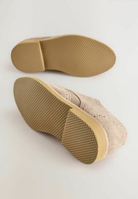 Chaussures en daim beige avec des accents perforés et une semelle en caoutchouc texturée. La semelle présente un motif en nid d'abeille pour une meilleure adhérence et durabilité.