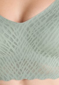 Detailný záber na zelenú textúrovanú látku s geometrickým vzorom na odeve, ktorý zobrazuje časť hrude a oblasti dekoltu.