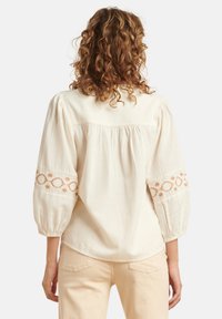 Cremfarbene Bluse mit Puffärmeln, die mit gestickten Akzenten in Rosttönen an den Manschetten und einer Raffung im Rücken versehen ist.