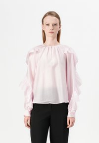 Blusa rosa chiaro con maniche a volantino, scollo rotondo e texture arricciata. Abbinata a pantaloni neri. Tessuto liscio e morbido.