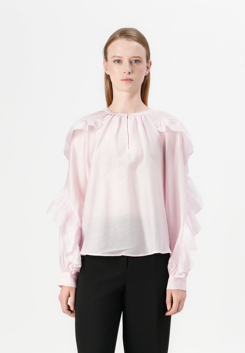 Blusa rosa chiaro con maniche a volantino, scollo rotondo e texture arricciata. Abbinata a pantaloni neri. Tessuto liscio e morbido.