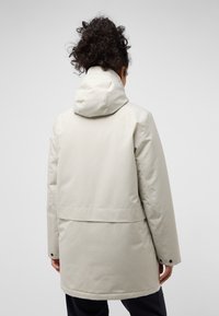 Veste à capuche beige clair avec une texture lisse, un dos légèrement plus long et des détails de bouton-pression aux poignets.