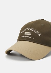 Olíva zöld sapka ívelt bézs viaszból. Hímzett "MONTPELLIER" és "DAYLIGHT STROLL" felirattal rendelkezik. Pamut anyag, hatpaneles kialakítás.